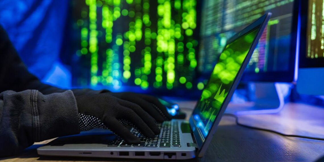 نمای نزدیک از دست‌های با دستکش سیاه در حال تایپ روی لپ‌تاپ با پس‌زمینه کدهای سبز رنگ (نماد هک).