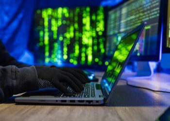 نمای نزدیک از دست‌های با دستکش سیاه در حال تایپ روی لپ‌تاپ با پس‌زمینه کدهای سبز رنگ (نماد هک).