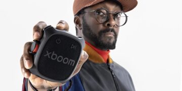 مردی با کلاه لبهدار و عینک که یک اسپیکر کوچک و مربعی مشکی برند XBOOM را به سمت دوربین نگه داشته است.