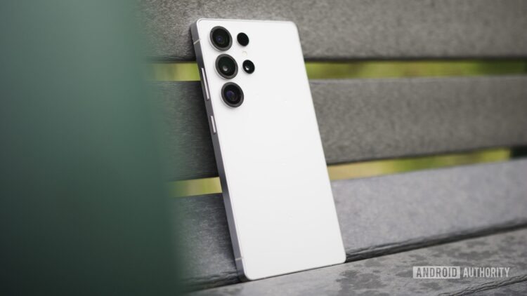 نمای نزدیک از پشت یک گوشی هوشمند سفید رنگ سری سامسونگ گلکسی S (احتمالاً S26 Ultra) که به یک سطح نیمکتی خاکستری متمایل شده و سیستم دوربین آن مشخص است.