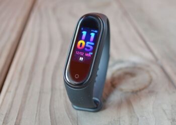 دست‌بند هوشمند شیائومی با نمایشگر باریک رنگی، نمایش ساعت و تاریخ روی صفحه، با بند مشکی سیلیکونی، قرارگرفته روی سطح چوبی با نور طبیعی