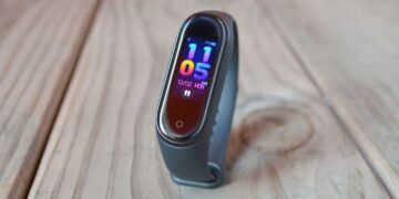 دستبند هوشمند شیائومی با نمایشگر باریک رنگی، نمایش ساعت و تاریخ روی صفحه، با بند مشکی سیلیکونی، قرارگرفته روی سطح چوبی با نور طبیعی