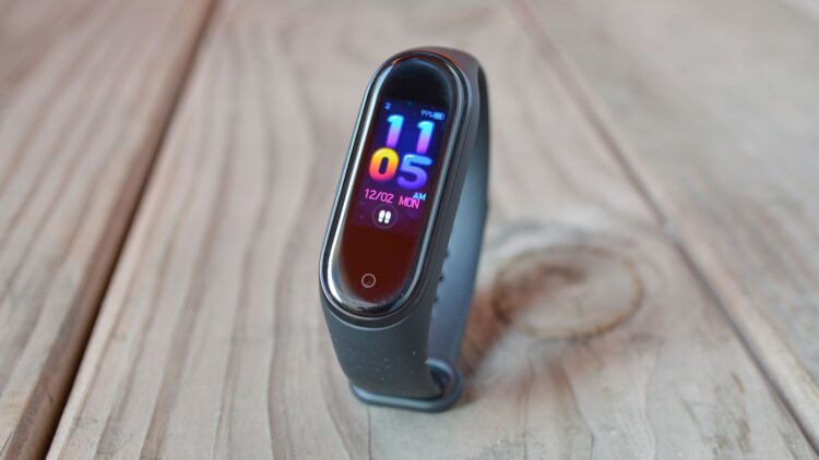 دستبند هوشمند شیائومی با نمایشگر باریک رنگی، نمایش ساعت و تاریخ روی صفحه، با بند مشکی سیلیکونی، قرارگرفته روی سطح چوبی با نور طبیعی