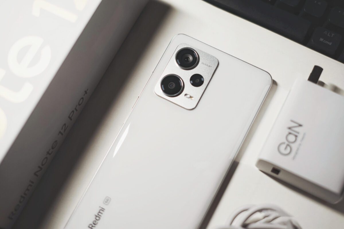 نمای تخت از پشت گوشی سفید رنگ ردمی نوت ۱۲ (نسخه پرو پلاس) که لنزهای دوربین و شارژر آن روی میز دیده میشود.