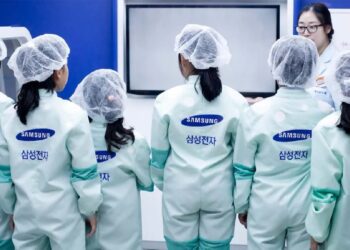 صفی از کارکنان یا بازدیدکنندگان با لباس‌های محافظ مخصوص (Cleanroom suit) و لوگوی سامسونگ.