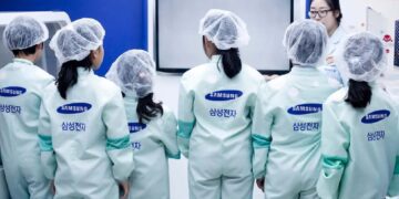 صفی از کارکنان یا بازدیدکنندگان با لباس‌های محافظ مخصوص (Cleanroom suit) و لوگوی سامسونگ.