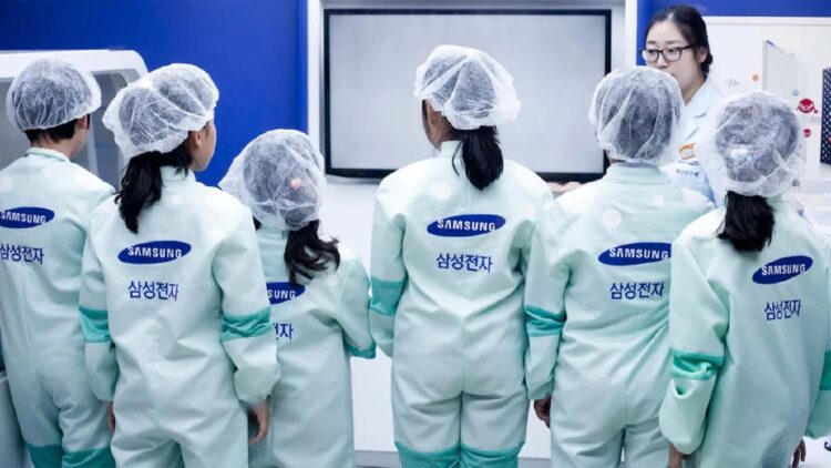 صفی از کارکنان یا بازدیدکنندگان با لباس‌های محافظ مخصوص (Cleanroom suit) و لوگوی سامسونگ.