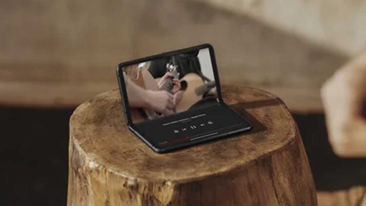 گوشی تاشوی سامسونگ در حالت نیمهباز روی سطح چوبی، در حال پخش ویدیو موسیقی روی نمایشگر داخلی، با تمرکز بر طراحی لولا و فرم استفاده رومیزی