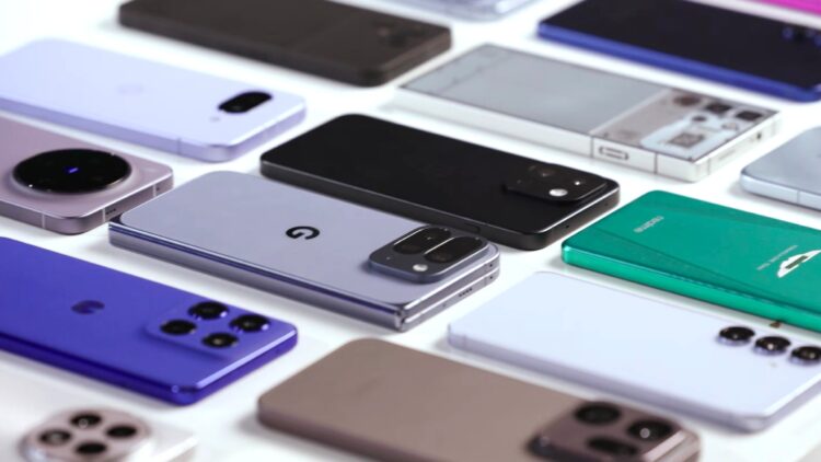 نمای نزدیک از ده‌ها گوشی هوشمند پرچمدار و میان‌رده سال ۲۰۲۵ که به صورت منظم روی میز سفید چیده شده‌اند.