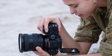 زنی در حال عکاسی یا فیلم‌برداری با دوربین سونی روی شن‌های ساحل با استفاده از صفحه نمایش دوربین.