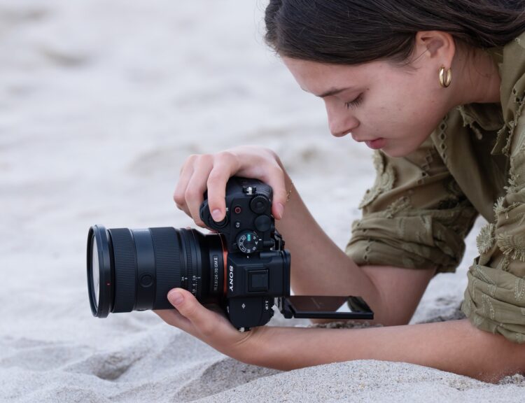 زنی در حال عکاسی یا فیلمبرداری با دوربین سونی روی شنهای ساحل با استفاده از صفحه نمایش دوربین.