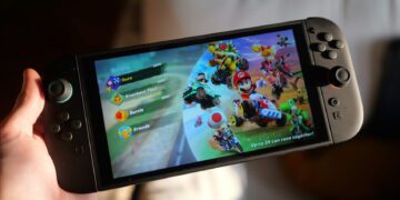 کنسول Nintendo Switch در حالت رومیزی روی میز کار، با نمایش بازی Mario Kart روی صفحهنمایش و دو کنترلر Joy‑Con جداشده در اطراف دستگاه