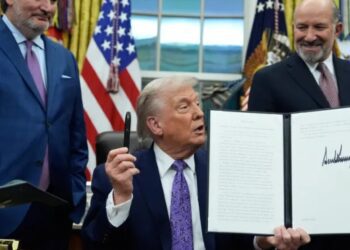 دونالد ترامپ در یک مراسم عمومی و روی صحنه، سند امضا شده‌ای را رو به دوربین نگه داشته است.