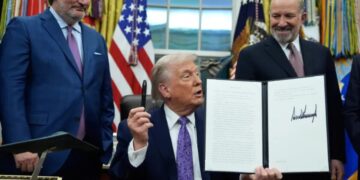 دونالد ترامپ در یک مراسم عمومی و روی صحنه، سند امضا شدهای را رو به دوربین نگه داشته است.