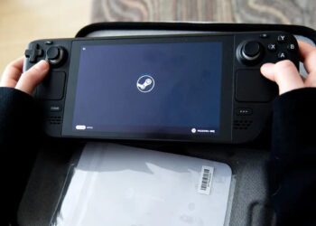 نمای نزدیک کنسول دستی Steam Deck با نمایش لوگوی Steam روی صفحه‌نمایش، طراحی کنترلرهای یکپارچه، آنالوگ‌استیک‌ها و دکمه‌های کنترلی، قرارگرفته روی سطحی نرم در محیط داخلی