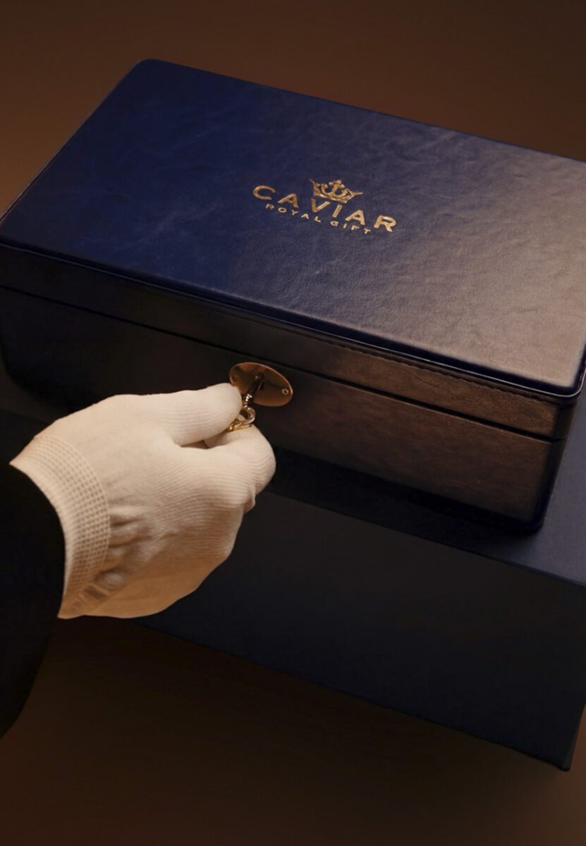 جعبه لوکس آبی‌رنگ کاویار با لوگوی طلایی برند که یک دستکش سفید، کلید طلایی را برای باز کردن آن در دست دارد