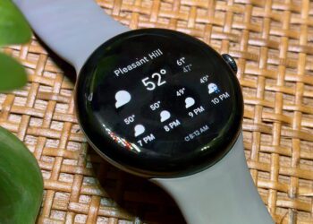 ساعت هوشمند با نمایش اطلاعات آب‌وهوا روی صفحه شامل دما و پیش‌بینی ساعتی، قرارگرفته روی سطح حصیری با بند سیلیکونی خاکستری، مناسب محتوای مرتبط با گجت‌های پوشیدنی و سلامت دیجیتال