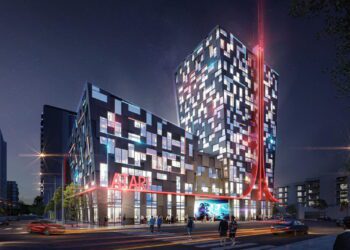 نمای شب از ساختمان هتل آتاری با معماری مدرن و نورپردازی ال‌ای‌دی قرمز، لوگوی Atari روی نمای خارجی، نمایش جلوه‌های فیوچریستیک و حضور خودروها و عابران در مقابل ورودی، مرتبط با پروژه هتل و مرکز سرگرمی آتاری در شهر