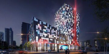 نمای شب از ساختمان هتل آتاری با معماری مدرن و نورپردازی ال‌ای‌دی قرمز، لوگوی Atari روی نمای خارجی، نمایش جلوه‌های فیوچریستیک و حضور خودروها و عابران در مقابل ورودی، مرتبط با پروژه هتل و مرکز سرگرمی آتاری در شهر