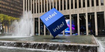 المان تبلیغاتی مکعب آبی Intel نصب‌شده در مرکز یک آب‌نمای شهری مقابل ساختمان اداری مدرن؛ تأکید بر برندینگ اینتل، معماری شهری و هویت بصری شرکت فناوری.