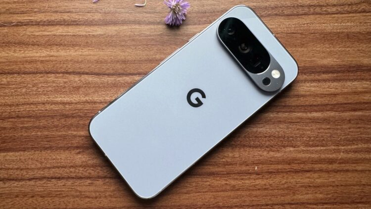 نمای پشت یک گوشی گوگل پیکسل (Google Pixel) با رنگ روشن که بر روی یک میز چوبی در کنار یک گل بنفش کوچک قرار دارد.