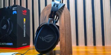 هدست ایسوس ROG Kithara قرار گرفته بر روی یک پایه چوبی عمودی در کنار جعبه محصول با پس‌زمینه دیواره آکوستیک.