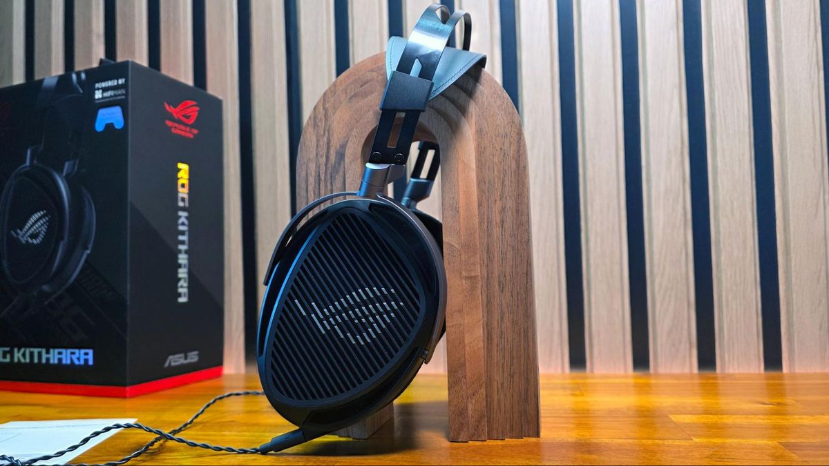 هدست ایسوس ROG Kithara قرار گرفته بر روی یک پایه چوبی عمودی در کنار جعبه محصول با پس‌زمینه دیواره آکوستیک.