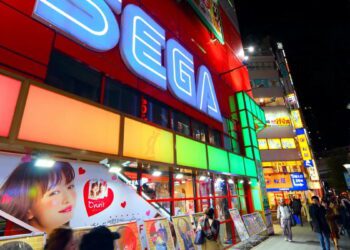 نمای شبانه از یک خیابان تجاری با ساختمان قرمز‌رنگ و تابلوی نورانی بزرگ SEGA، ویترین فروشگاه‌های بازی، پوسترهای تبلیغاتی رنگی و تابلوهای نئونی متعدد در محیط شهری پرنور