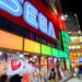 نمای شبانه از یک خیابان تجاری با ساختمان قرمز‌رنگ و تابلوی نورانی بزرگ SEGA، ویترین فروشگاه‌های بازی، پوسترهای تبلیغاتی رنگی و تابلوهای نئونی متعدد در محیط شهری پرنور