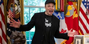 ایلان ماسک با کلاه "DOGE" و کت مشکی در مقابل پرچم‌های ایالات متحده در حال صحبت با هیجان.