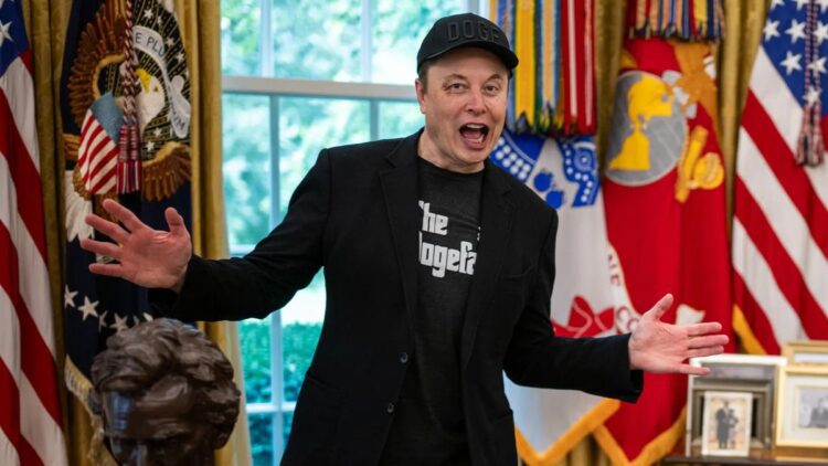 ایلان ماسک با کلاه "DOGE" و کت مشکی در مقابل پرچمهای ایالات متحده در حال صحبت با هیجان.