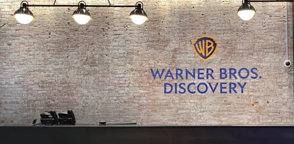 نمای عریض از دیوار آجری داخلی با لوگوى Warner Bros. Discovery در مرکز، شامل نشان WB زرد‑آبی و تایپوگرافی Warner Bros. Discovery؛ نورپردازی صنعتی سقفی و پیشخوان تیره در پایین تصویر.