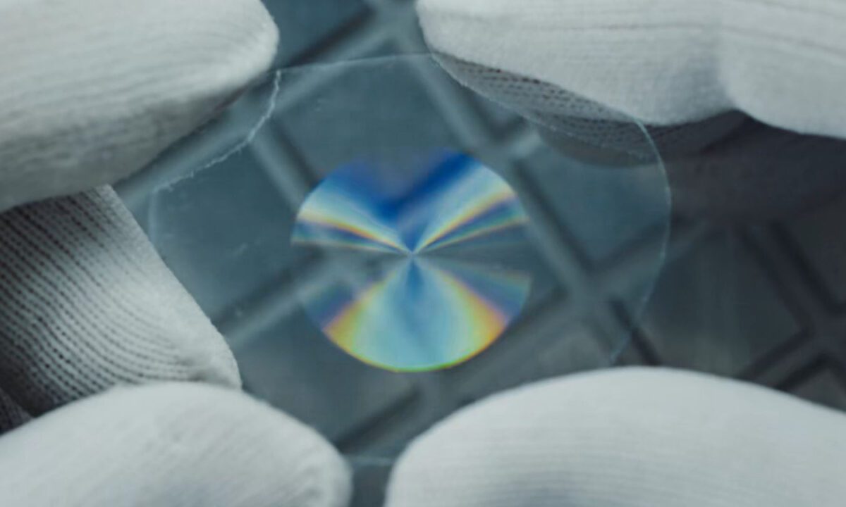 لنز عینک هوشمند IXI که توسط دستکش‌های محافظ سفید نگه داشته شده و شکست نور رنگین‌کمانی را در مرکز نشان می‌دهد.