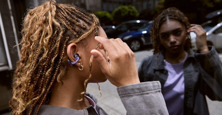 زنی با موهای بافته شده که یک هدفون بنفش JBL Sense را به گوش دارد و در فضای شهری ایستاده است.