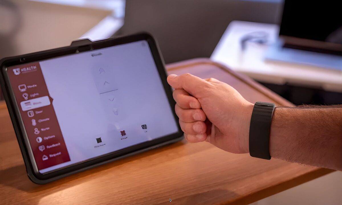 نمای نزدیک از دست فردی که یک مچ‌بند مشکی هوشمند به دست دارد و با حرکات انگشت در حال کنترل محتوای روی تبلت مقابل خود است.