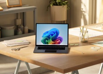 یک لپ‌تاپ باز روی میز کار چوبی که والپیپر آبی ویندوز ۱۱ را نشان می‌دهد و نوری ملایم از پنجره به آن می‌تابد.