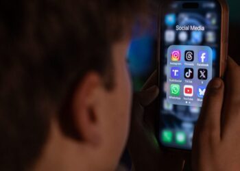 نمای نزدیک از نیم‌رخ یک نوجوان که در تاریکی به صفحه‌نمایش گوشی خود و آیکون‌های برنامه‌های شبکه‌های اجتماعی نگاه می‌کند.