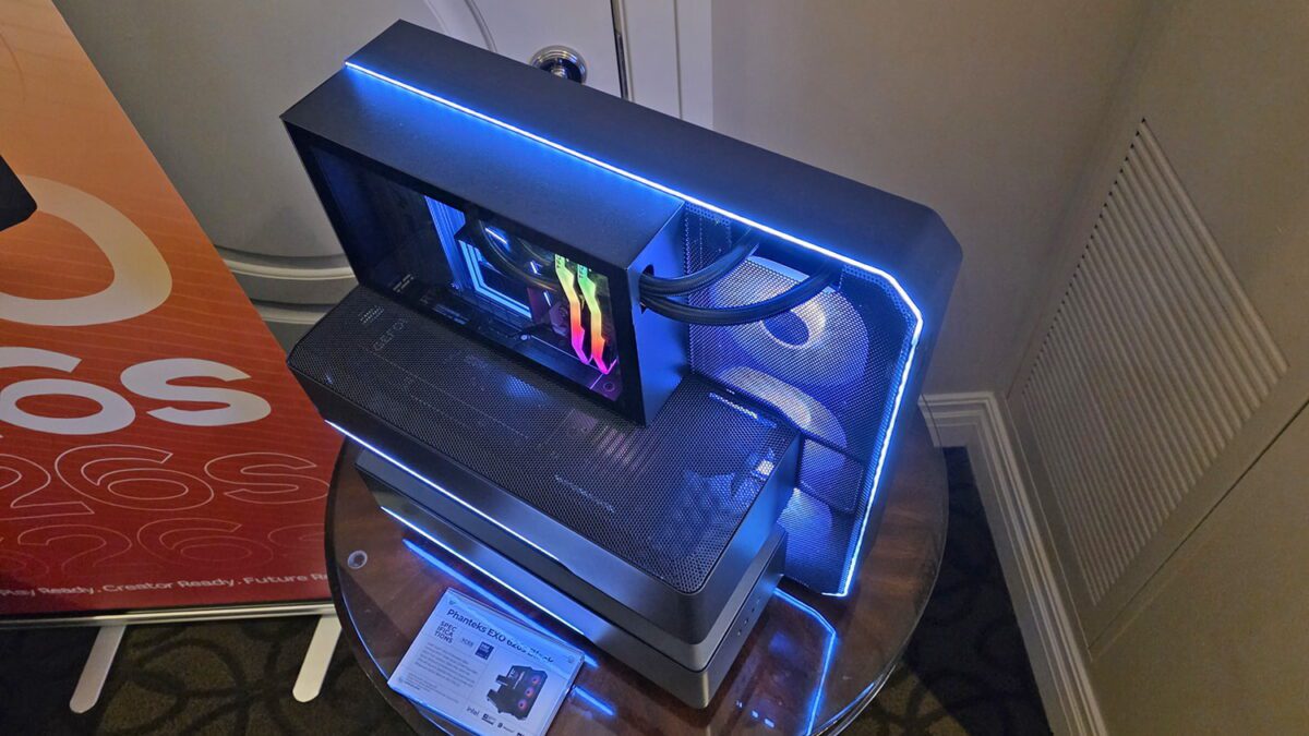 نمای بالا از یک کیس کامپیوتر مفهومی با طراحی ماژولار لایه‌ای، شامل پنل‌های مشکی مات، نورپردازی خطی LED آبی، پنل شیشه‌ای شفاف برای نمایش قطعات داخلی و فن‌های RGB؛ قرارگرفته روی میز نمایشگاهی با کارت مشخصات محصول.