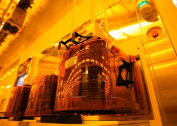 نمای داخلی یک کارخانه تولید نیمههادی (Cleanroom) با حاملهای خودکار ویفر FOUP شفاف نارنجی روی خطوط تجهیزات؛ نورپردازی زرد صنعتی و تمرکز بر فرآیندهای خودکار جابهجایی و نگهداری ویفر سیلیکونی.