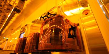 نمای داخلی یک کارخانه تولید نیمه‌هادی (Cleanroom) با حامل‌های خودکار ویفر FOUP شفاف نارنجی روی خطوط تجهیزات؛ نورپردازی زرد صنعتی و تمرکز بر فرآیندهای خودکار جابه‌جایی و نگه‌داری ویفر سیلیکونی.