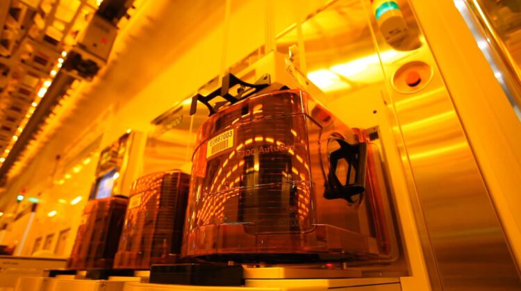 نمای داخلی یک کارخانه تولید نیمه‌هادی (Cleanroom) با حامل‌های خودکار ویفر FOUP شفاف نارنجی روی خطوط تجهیزات؛ نورپردازی زرد صنعتی و تمرکز بر فرآیندهای خودکار جابه‌جایی و نگه‌داری ویفر سیلیکونی.