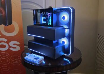 نمای سه‌رخ از کیس کامپیوتر پیشرفته با ساختار چندبخشی پله‌ای، پنل جلویی مشبک با نورپردازی LED آبی، سه فن خنک‌کننده دایره‌ای، پنل شیشه‌ای کناری و نمایش ماژول سخت‌افزاری داخلی در محیط نمایشگاهی.