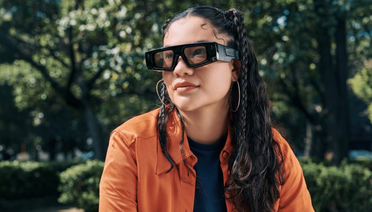 نمای محیطی از عینک هوشمند Snapchat Spectacles در فضای باز، با فریم مشکی و لنزهای زاویه‌دار که طراحی آن بر استفاده از واقعیت افزوده و تعامل دیجیتال در محیط طبیعی تأکید دارد؛ پس‌زمینه شامل پوشش گیاهی و نور طبیعی است.