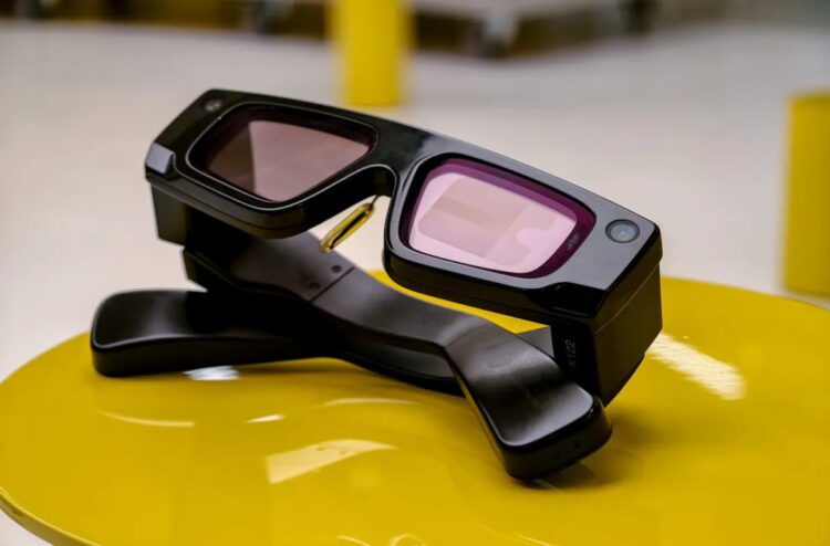 نمای استودیویی عینک هوشمند Snapchat Spectacles با طراحی ضخیم و صنعتی، لنزهای رنگی مایل به بنفش، فریم مشکی مات و ماژول‌های دوربین یکپارچه در گوشه‌های فریم؛ محصول روی پایه‌ی نگهدارنده‌ی مشکی و سطح زرد براق قرار گرفته است.