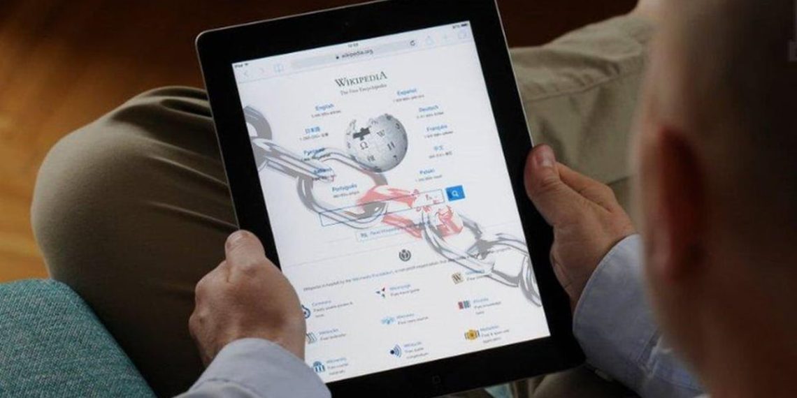 فردی در حال استفاده از تبلت که صفحه اصلی ویکی‌پدیا روی آن نمایش داده شده و نمادی از دسترسی آنلاین به دانش، یادگیری دیجیتال و محتوای مشارکتی است
