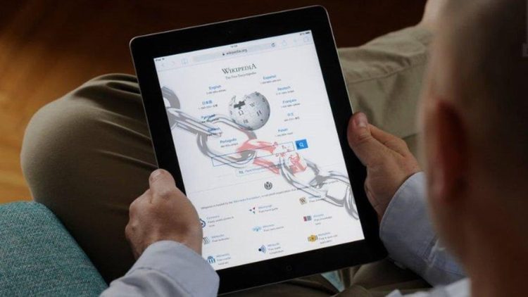 فردی در حال استفاده از تبلت که صفحه اصلی ویکی‌پدیا روی آن نمایش داده شده و نمادی از دسترسی آنلاین به دانش، یادگیری دیجیتال و محتوای مشارکتی است