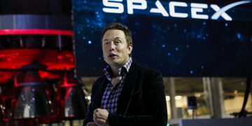 تصویر یک رویداد مرتبط با صنعت فضایی و فناوری که در آن ایلان ماسک با پوشش نیمه‌رسمی در فضای داخلی مدرن ایستاده است. در پس‌زمینه، لوگوی بزرگ SpaceX و سازه‌ای صنعتی با نورپردازی قرمز و آبی دیده می‌شود که تداعی‌گر محیط مهندسی، پرتاب فضایی و نوآوری در فناوری هوافضا است.