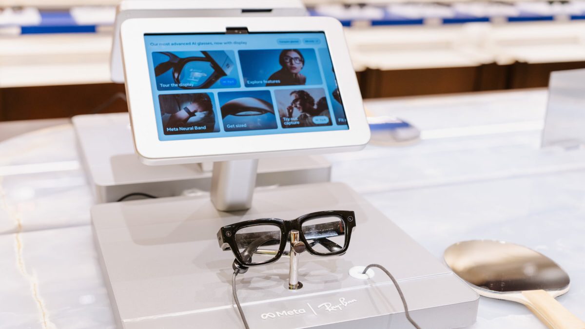 نمای نزدیک از عینک هوشمند Meta Ray‑Ban روی پایه نمایش، در مقابل نمایشگری لمسی که گزینه‌هایی مانند Explore features، Meta Neural Band و Tour the display را نشان می‌دهد؛ تمرکز تصویر بر رابط کاربری و سخت‌افزار محصول است.