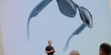 صحنه معرفی عینک هوشمند Meta Ray‑Ban روی استیج، با تصویر بزرگ عینک در پس‌زمینه؛ طراحی کلاسیک Ray‑Ban با فریم تیره، لنزهای دودی و ماژول‌های الکترونیکی یکپارچه در دسته‌ها نمایش داده شده است.