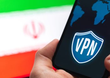 دستی که گوشی هوشمندی با نماد VPN و نقشه جهان را در مقابل پرچم ایران نگه داشته است.